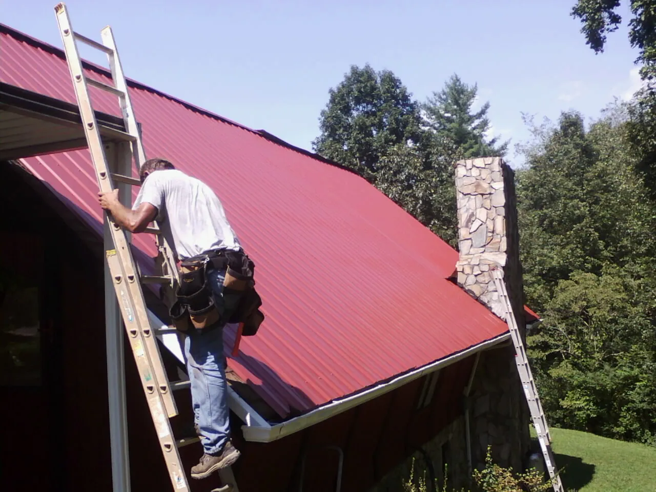 Roofer on a ladder beside a red metal roof and stone chimney — Soffit & Fascia Repair by Roofing Brothers in Fairchance